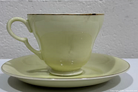 Side view of cream porcelain tea cup on saucer showing heart-shaped handle and gold rim