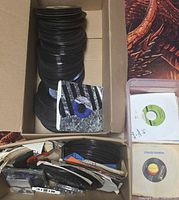 Box containing large stack of 45 rpm records, sleeves, cassette tapes visible