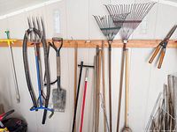 Wall-mounted view of pitchfork, spade, two leaf rakes, hedge shears, weed puller