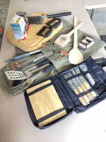 All items spread on table: stainless tools, wooden utensils, thermometer boxes, apron, board, case