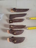 Six brown leather knife sheaths laid out showing tooling, snap buttons, and varying lengths