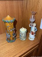 Three items on shelf: tall carved wax candle, shorter candle in ceramic holder, figural lamp