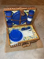 Open wicker picnic basket showing included blue plastic plates, tumblers, cutlery, napkins