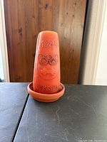 Front view of orange wax candle showing 1964 Innsbruck Olympic design seated in terracotta saucer
