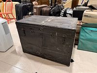 Front and right side view of black metal steamer trunk showing riveted panels, latches, missing left handle