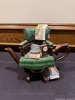 Front view of teapot showing chair form, spout, handle, rum bottle, map