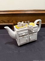 Front view of ceramic kitchen sink teapot showing spout, handle, cabinet detailing and dish rack