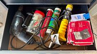 Overview of drawer contents showing drill, yellow tool, grease cartridges, hardware jar, boxed multimeters