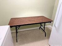 Full view of brown laminate folding table with black legs against wall