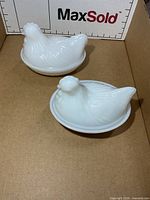 Both milk glass hen-on-nest dishes with lids in cardboard photo booth