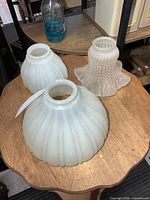 Three glass lamp shades on tabletop: two white ribbed milk glass, one frosted hobnail ruffled