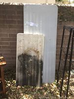 Two plastic folding leg tables side by side leaning against a brick wall. Large table is clean and white, small table has heavy black stains and discoloration.