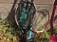 Two tennis rackets standing against wall, black and red color, with a tennis racket cover branded Head partially visible