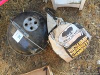Black round Weber charcoal grill lid with vent holes and bag of 100% lump Mesquite charcoal beside it on the ground.