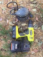 Top view of Ryobi cordless drill with battery attached, battery charger with power cord, and blue electric palm sander on grass and leaves background.