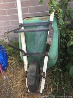 Full view of wheelbarrow frame showing green basin, white handles, black metal support bracket and single tire with visible wear and rust.