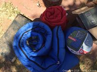 Two rolled sleeping bags, one blue and one red, plus a new unopened Embark brand sleeping bag in plastic packaging.