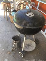 Wide view of large black Weber kettle grill with lid and vent holes on porch floor, accompanied by metal chimney starter and attached ash catcher pan below.