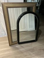 Rectangular bronze-tone framed mirror and arched black mirror standing together