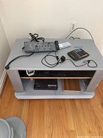 Gray two-tier TV stand holding surge protector, calculator, USB cables