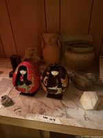 Lot overview showing two Japanese dolls, terracotta jar, beige urn, gravy boat, marble paperweight, mineral specimen