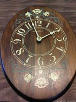 Front view of oval Caravelle wood wall clock showing dial, numerals and floral motifs