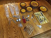 Overall lot view on wooden table: glass tumblers, ceramic dinnerware stacks, flatware, kitchen tools