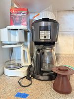 Two Black & Decker coffee makers side by side with carafes, Melitta cone box and loose brown plastic part visible