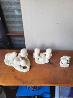 Three white/ivory resin figurines on shelf: elephant with sheep group, children group, small gift box with bear