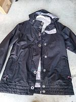 Full view of black hooded jacket laid flat showing front, pockets, snaps, zipper, hood