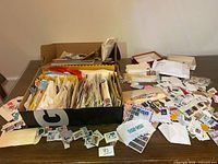 Cardboard box filled with envelopes containing stamps, loose stamps spread on table