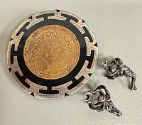 Front view of brooch with Aztec calendar design alongside two sterling charms
