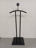 Front view of black metal valet stand showing circular head, curved shoulder bar and base