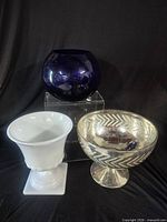 Three vessels grouped: cobalt blue round vase on acrylic riser, white milk glass pedestal vase, silver mercury glass pedestal bowl