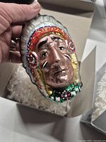 Front view of hand holding the glass chief ornament over box with packing