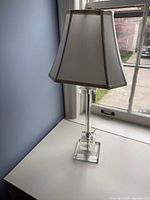 full view of lamp on table beside window showing glass base and white shade
