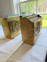 Two gold-tone embossed magazine holders on table showing decorative panels and contents for scale