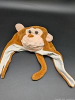 Front view of brown plush monkey hat with ear flaps and tail extension