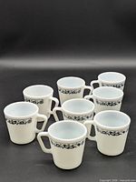 Eight matching white Pyrex mugs with blue floral bands grouped on black background