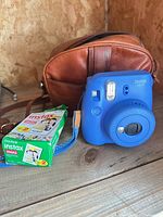Blue Fujifilm Instax Mini 9 camera, brown case, box of Instax Mini film displayed on wooden surface
