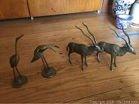 Photo showing two brass crane figurines with elongated necks and two brass antelope figurines with long twisted horns on a wooden surface.