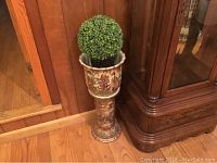 Full view of ceramic jardiniere with detailed Asian inspired paintings and plastic topiary inside, standing on hardwood floor next to wood cabinet.