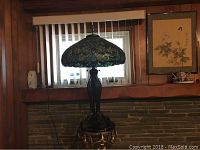 Full view of the Tiffany style stained glass lamp with metal base on a small ornate side table against a window with vertical blinds.