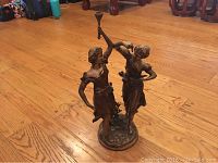Full view of the brass sculpture showing two women dancing with one holding a horn-like object, standing on an oval base with detailed foliage, on a wooden floor.