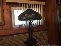 Full view of stained glass Tiffany style lamp on table showing metal base and stained glass shade with leaf floral design under window with blinds.