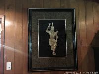 Full view of framed Asian wall art showing the hand-carved gold figure on black background within leaf-like patterned border and black frame.