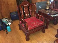 View of the entire wooden lounge chair showing carved arms and legs with mother of pearl inlay on backrest, and red silk seat cushion wrapped in plastic.
