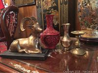 View of brass ram statue, carved red vase, smaller brass-tone vase, and brass candlestick holder on a wooden surface.