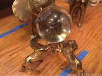 Photo showing two brass elephants in the background and a trio of brass horses holding a glass orb in the foreground on a wooden floor.