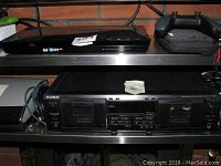Sony BDP-S5100 Blu-ray player positioned above a Sony TC-WE435 dual cassette deck on a shelf with some books and gaming controller nearby.